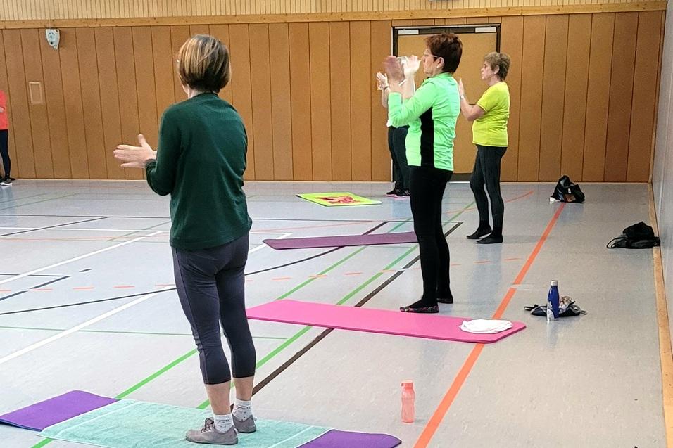 Gruppentraining in einer Halle, mit Teilnehmern auf Matten, die aktiv Übungen durchführen und applaudieren.