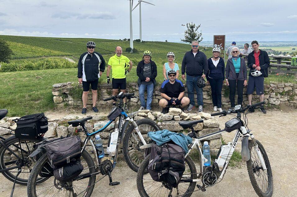 Gruppe von Radfahrern posiert vor Windrädern und grünem Landschaftshorizont, Fahrräder liegen vor ihnen am Boden.