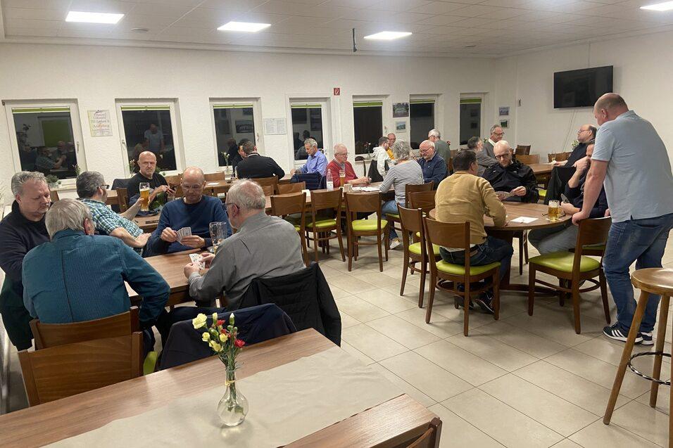 Gruppenszene in einem Versammlungsraum mit Männern, die an Tischen sitzen und sich unterhalten.