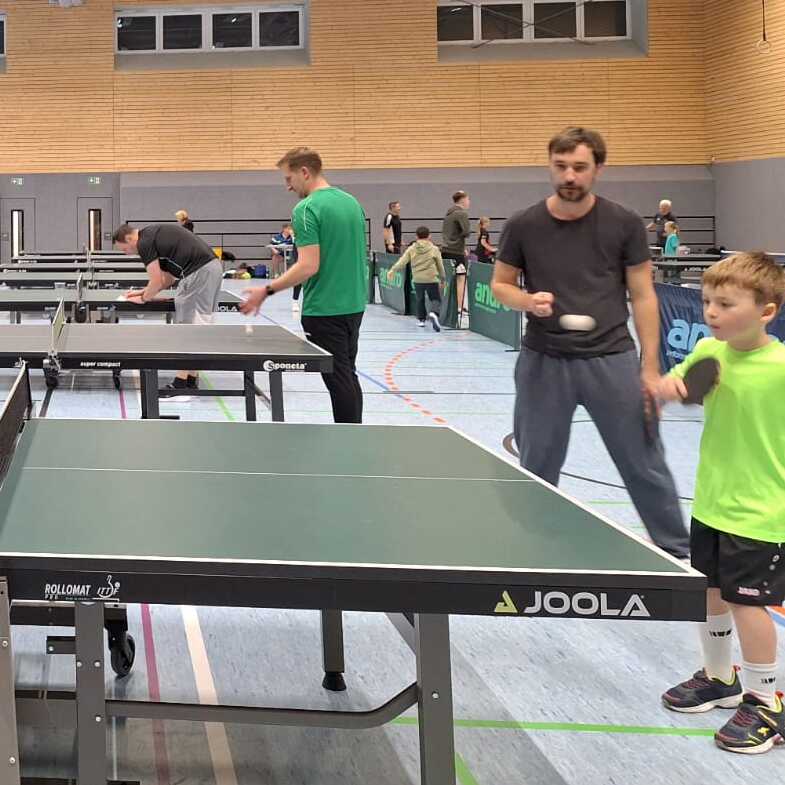 Gruppe von Spielern unterschiedlichen Alters beim Tischtennis in einer Sporthalle mit mehreren Tischtennisplatten.