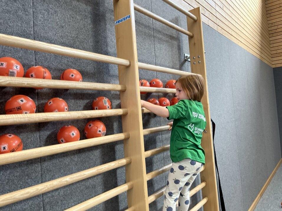 Kind in grünem T-Shirt klettert an einer Holzleiter, während orangefarbene Bälle in der Wandablage sind.
