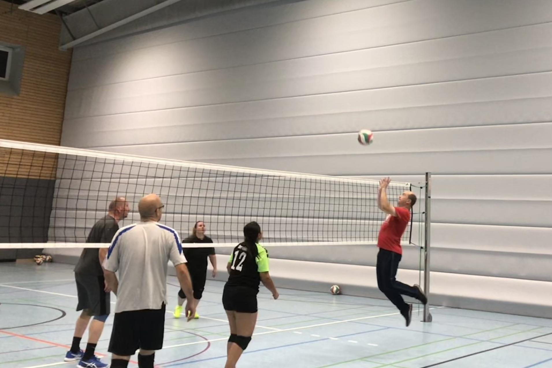 Volleyballspiel in einer Halle, ein Spieler springt zum Angriff, während andere warten. Netz und Ball sichtbar.
