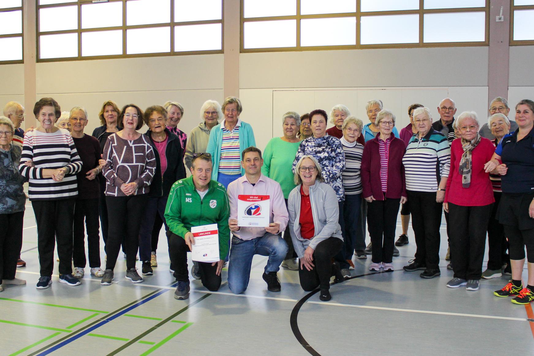 Gruppenfoto von Seniorinnen und Senioren in einem Sportraum, einige halten Preisurkunden, alle lächeln.